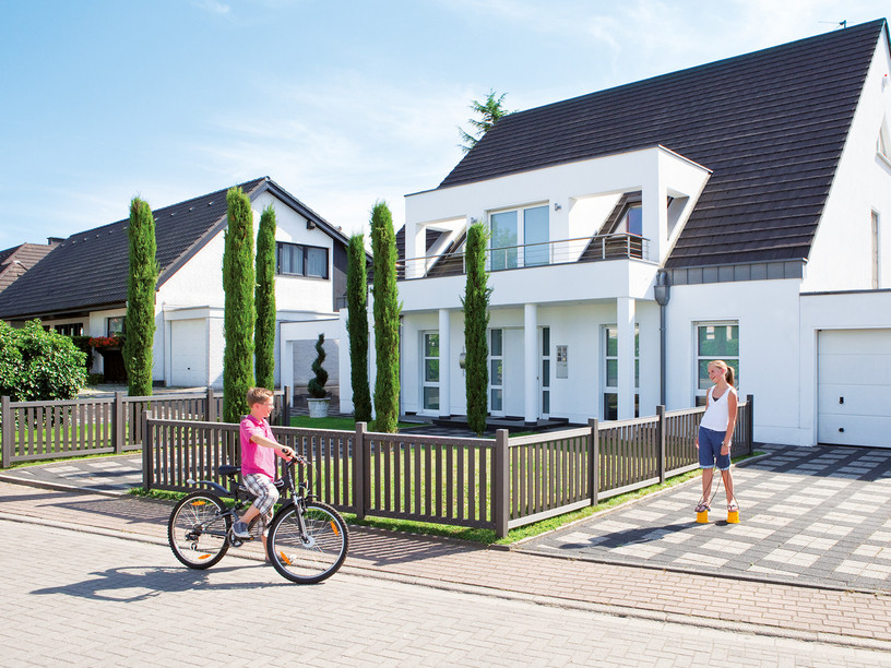 Modernes Einfamilienhaus mit Vorgartenzaun aus WPC, Kind mit Fahrrad und Frau im Einfahrtsbereich.