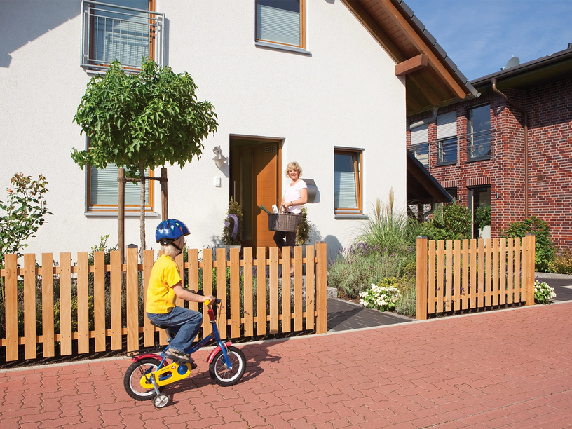 Holz-Vorgartenzaun vor Einfamilienhaus, spielendes Kind auf Fahrrad, Frau an der Haustür.