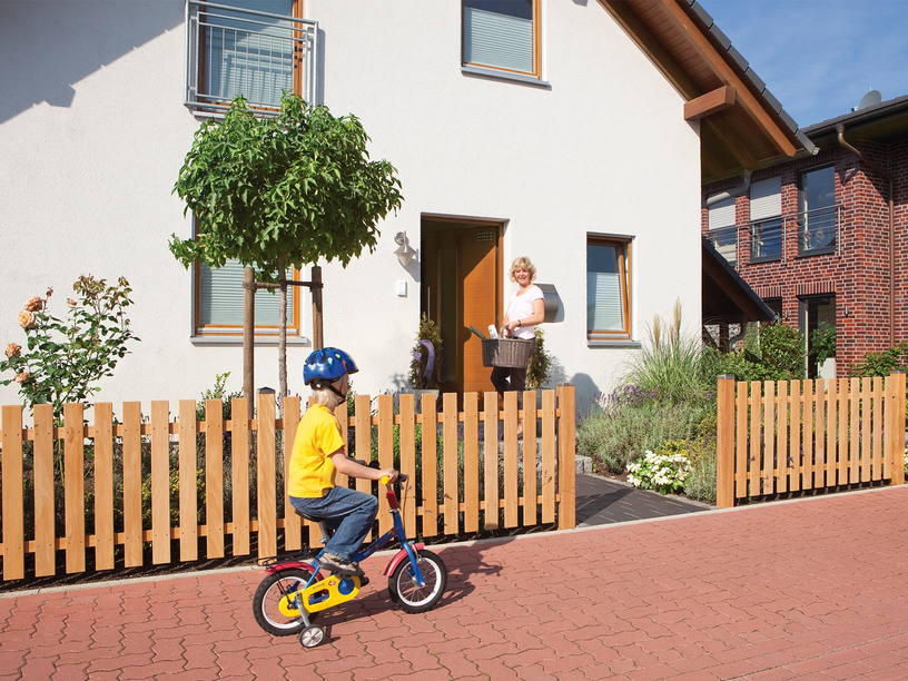 VorgartenzauHolz-Vorgartenzaun vor Einfamilienhaus, spielendes Kind auf Fahrrad, Frau an der Haustür.